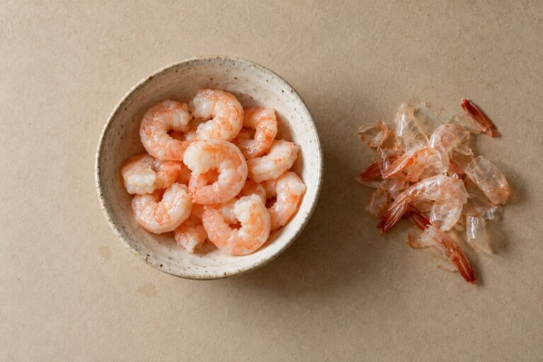 Bowl of cooked peeled shrimp pieces beside a small pile of discarded shrimp shells and tails